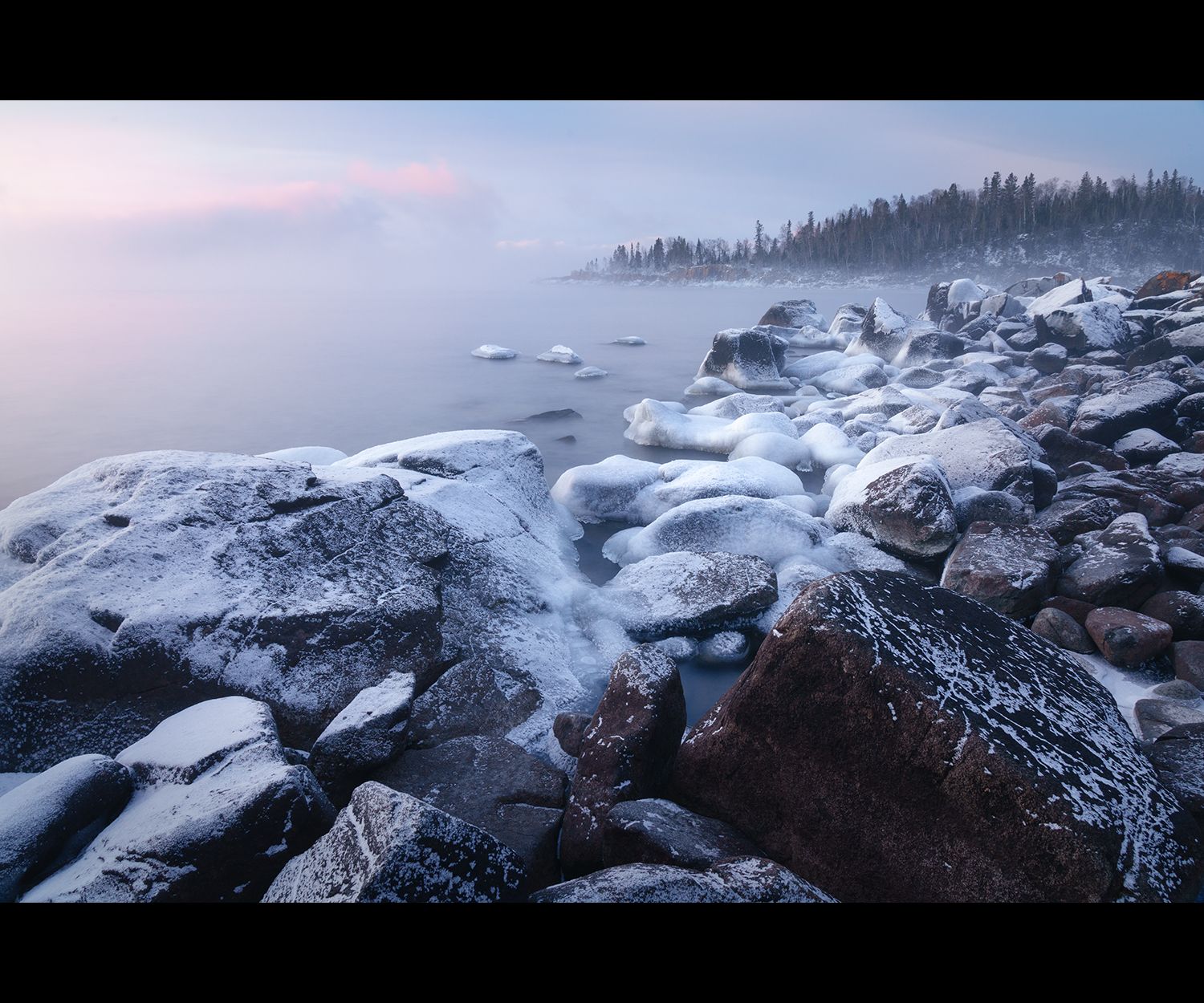 Rocky shoreline covered in frost before sunrise, with mist and soft pink clouds