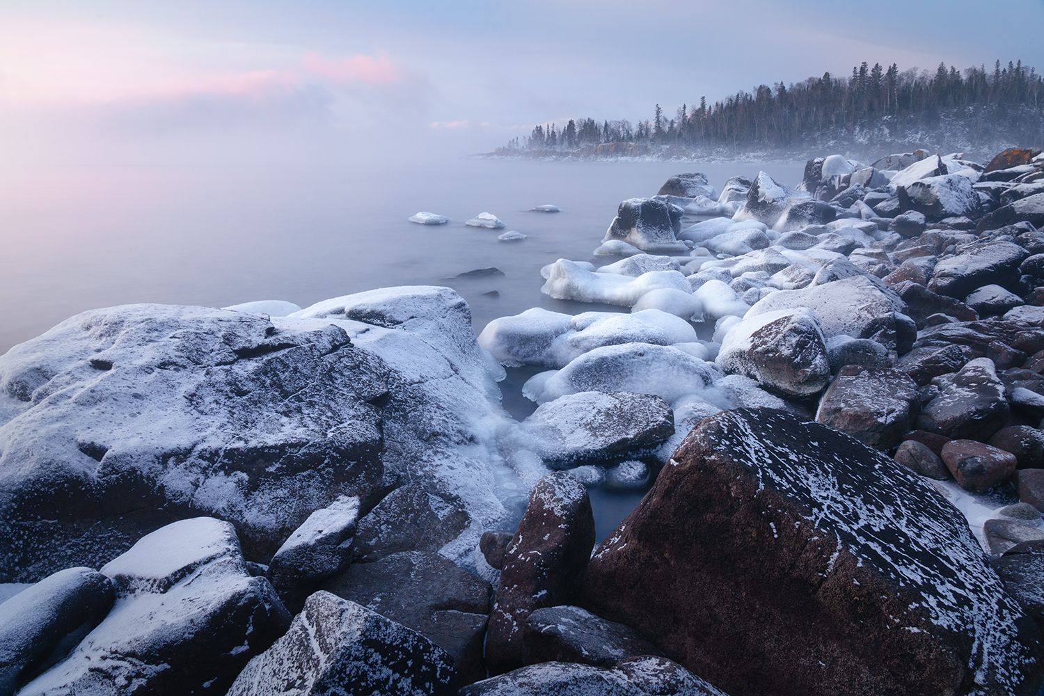 Rocky shoreline covered in frost before sunrise, with mist and soft pink clouds