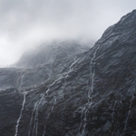 An ominous wall of mountains with waterfalls streaming down. A glacier is barely visible admist the clouds.