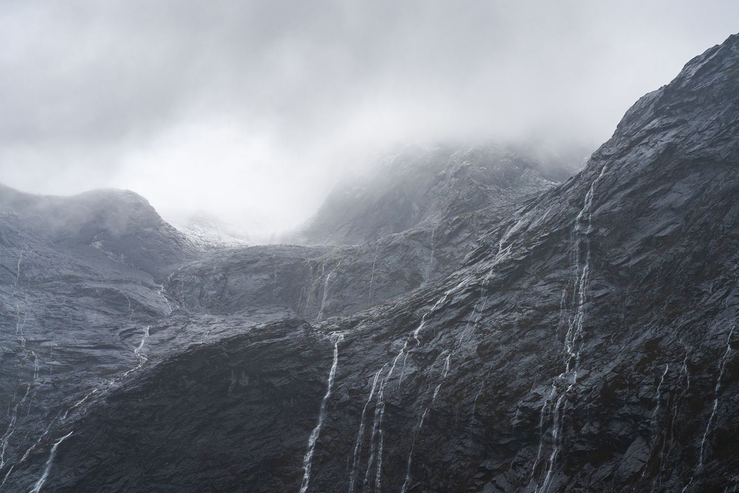 An ominous wall of mountains with waterfalls streaming down. A glacier is barely visible admist the clouds.